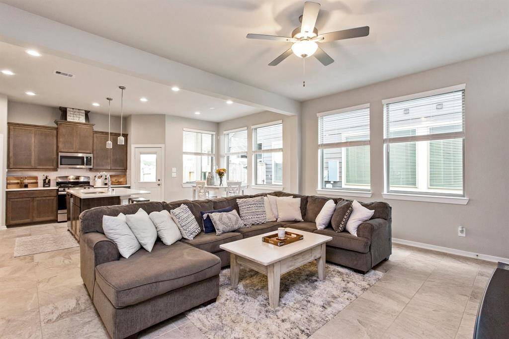 This spacious open-concept living area features a modern kitchen with dark wood cabinets and stainless steel appliances, a bright dining nook, and plenty of natural light from multiple large windows. The ceiling fan and neutral tones create a welcoming atmosphere.