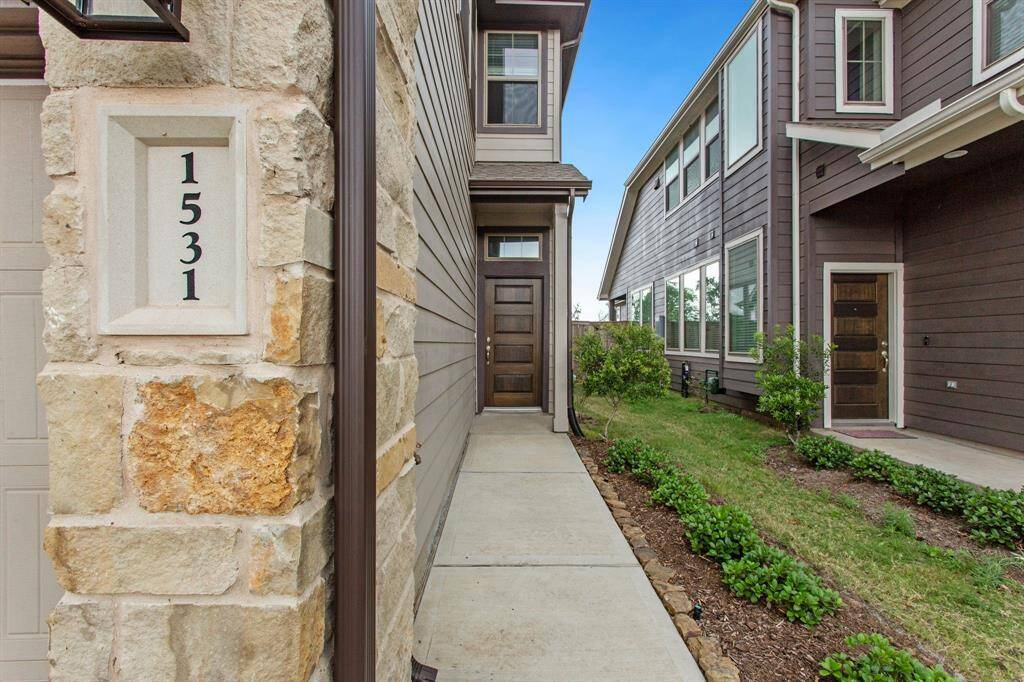 This photo shows the entrance of a modern townhouse with a stone accent wall featuring the number 1531. A narrow concrete path leads to a dark wooden door, flanked by well-maintained landscaping.