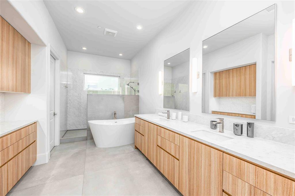 This bathroom features a modern design with a spacious layout. It includes a freestanding bathtub, large walk-in shower, and double vanity with wood cabinetry and sleek finishes. Bright and airy with ample natural light and minimalist decor.
