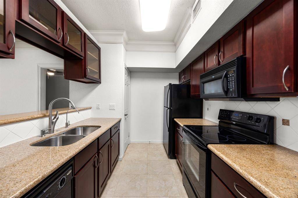 Stylish galley kitchen features rich wood cabinetry, granite countertops, black appliances, and a double sink with a modern faucet. Overlooks living room.