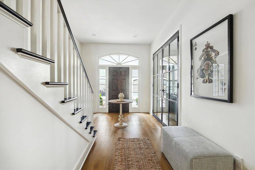 Step into this bright and sophisticated entryway that sets the tone for the rest of the home. The space features rich wood flooring and a striking staircase with contrasting black accents. Natural light streams through the arched transom window above the front door, creating a warm and welcoming ambiance.