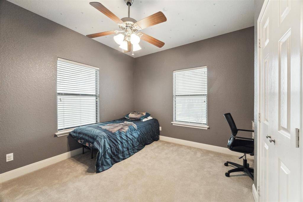 This is a modest-sized bedroom with neutral gray walls and carpeted flooring. It features two windows for natural light, a ceiling fan with lighting, and is currently furnished with a single bed and a desk chair. The room appears well-maintained and move-in ready.