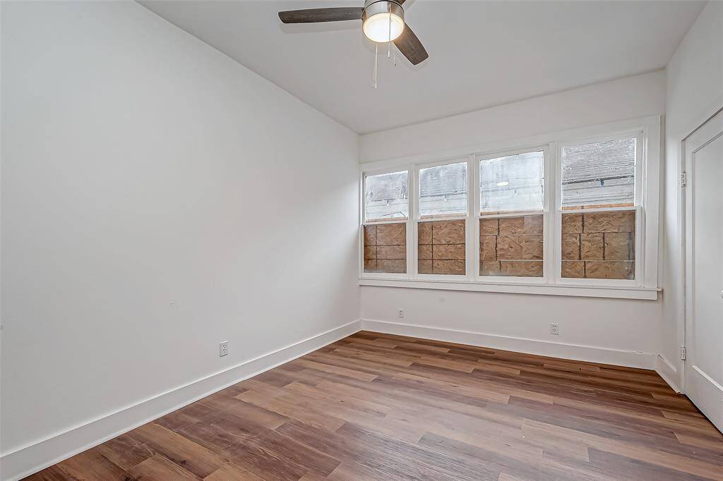 This well-lit bedroom features a full wall of windows, illuminating the fresh white walls and wood flooring. The ceiling fan’s light fixture adds a warm glow, complementing the natural light streaming in.
