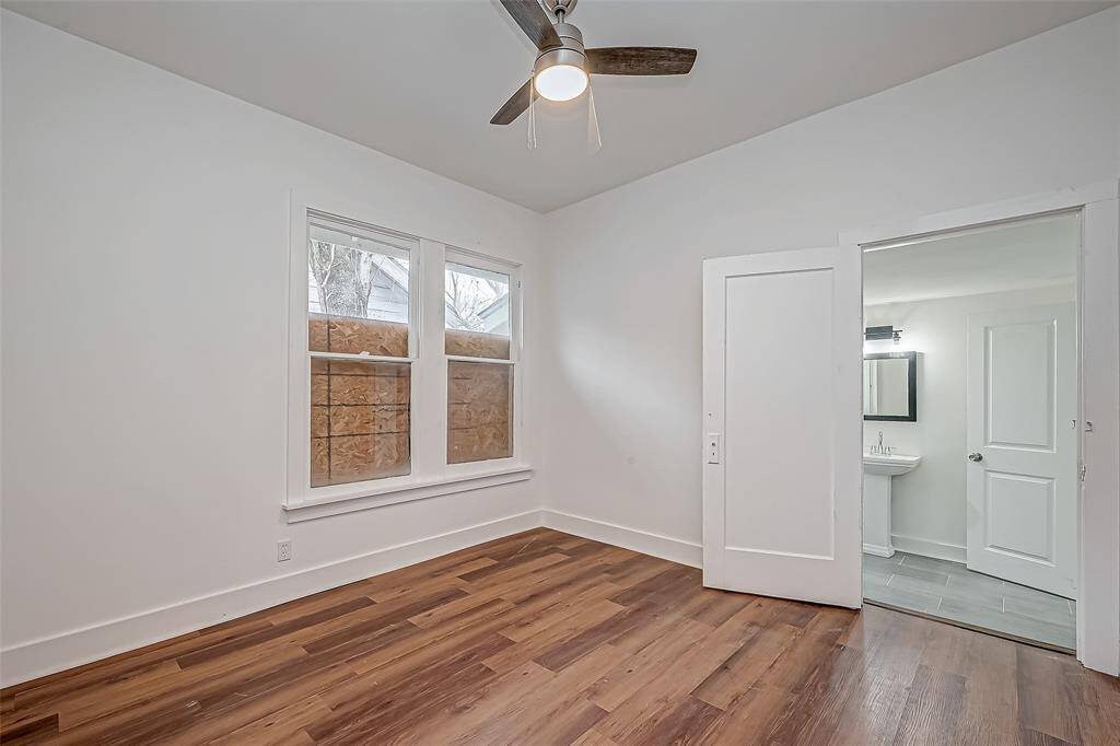 The primary bedroom offers generous space, continuing the home’s crisp aesthetic with white walls and warm wood flooring. The ceiling fan’s lighting fixture casts a soft glow, complementing the natural brightness from the window.