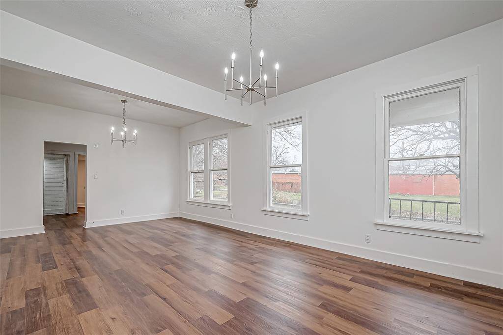 Defined yet connected, the dining area sits just beside the living room, subtly distinguished by a ceiling beam that creates visual separation. The white walls and ceiling enhance the sense of space, while the warm wood flooring adds depth. Twin chandeliers overhead complete the setting.
