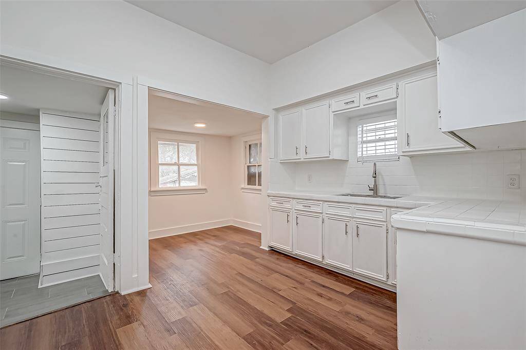 Through an open doorway, the kitchen continues the home’s crisp white aesthetic, complemented by shiny wood floors. All-white cabinetry and countertops maximize brightness, while a dedicated nook offers a cozy breakfast spot.