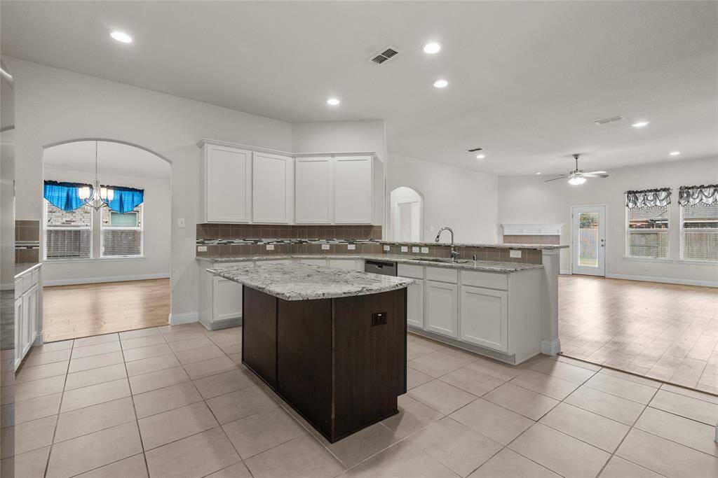 This spacious kitchen features white cabinetry, granite countertops, an island, modern appliances, tile flooring, and opens to a large, well-lit living area with ample natural light.  This large kitchen is the center of the home