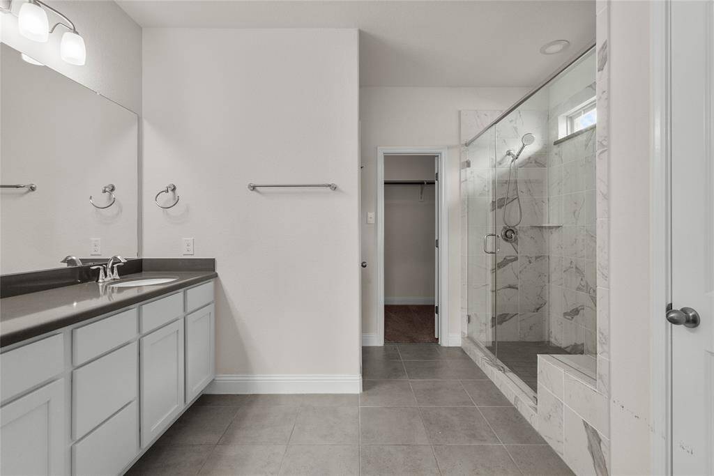 This is a modern bathroom featuring a double vanity with ample storage, spacious walk-in shower with clear glass doors. The room has tiled flooring and a neutral color scheme. A door at the back leads to the oversized primary closet