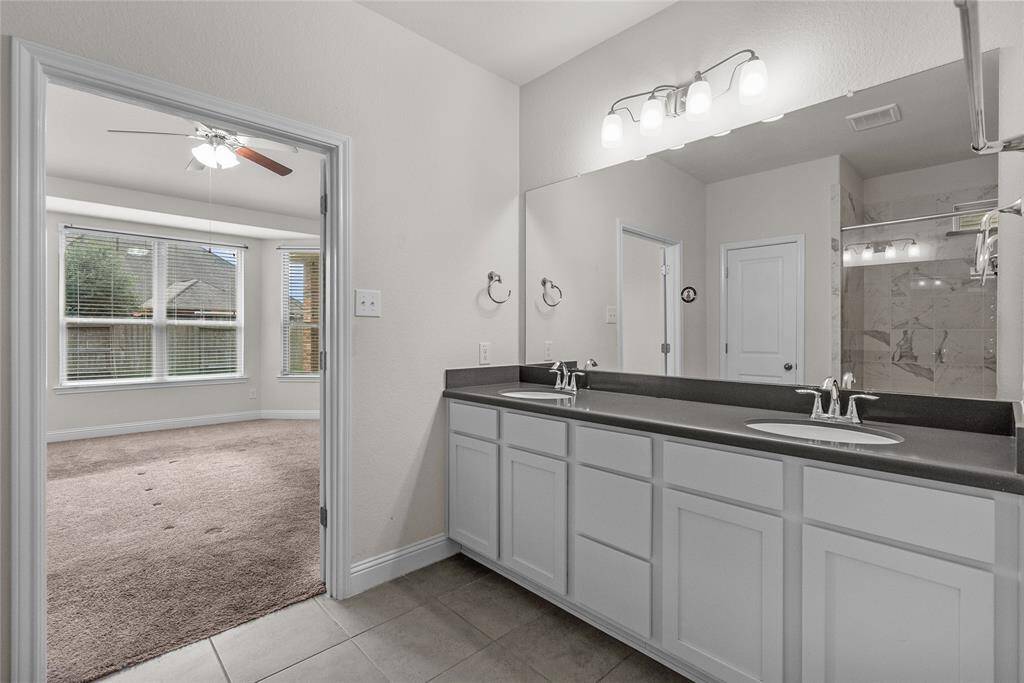 This is a well-lit, spacious en-suite bathroom featuring dual sinks, ample counter space, and a large mirror with modern lighting above. It opens to a primary bedroom with tons of windows that brings in natural light.