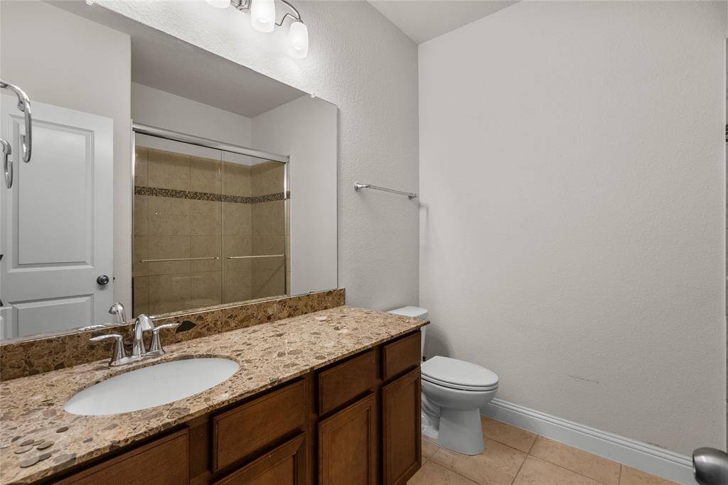 This is one of three bathrooms downstairs.  This room features a single vanity with ample counter space, under-sink cabinetry, a large mirror with overhead lighting, a walk-in shower with glass doors, and a toilet. The space has neutral tones and tiled flooring.