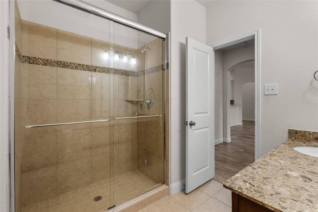 You're looking at a modern bathroom featuring a large walk-in shower with glass doors and decorative tiling, complemented by a granite vanity top. The space is well-lit and opens into a hallway leading to additional rooms.