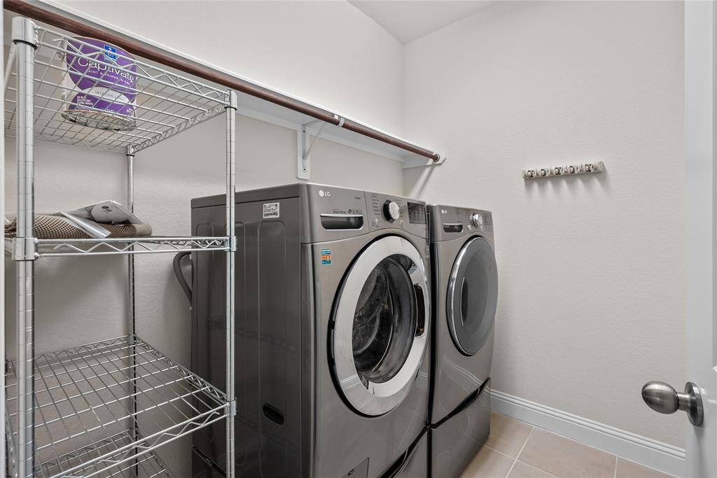 This is a neat and large laundry room featuring modern, front-loading washer and dryer units, ample shelving for storage, and a hanging rack for convenience. The room is finished with tile flooring and a clean, neutral color scheme.