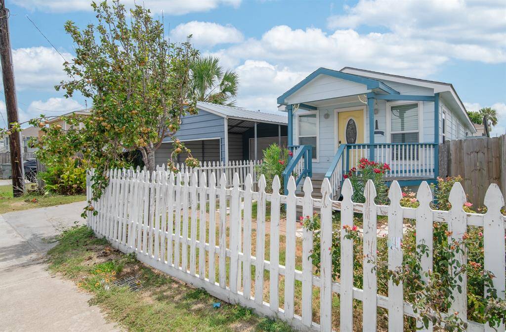 Adorned with trees, a covered parking area, and a covered porch, the property is surrounded by a white picket fence.