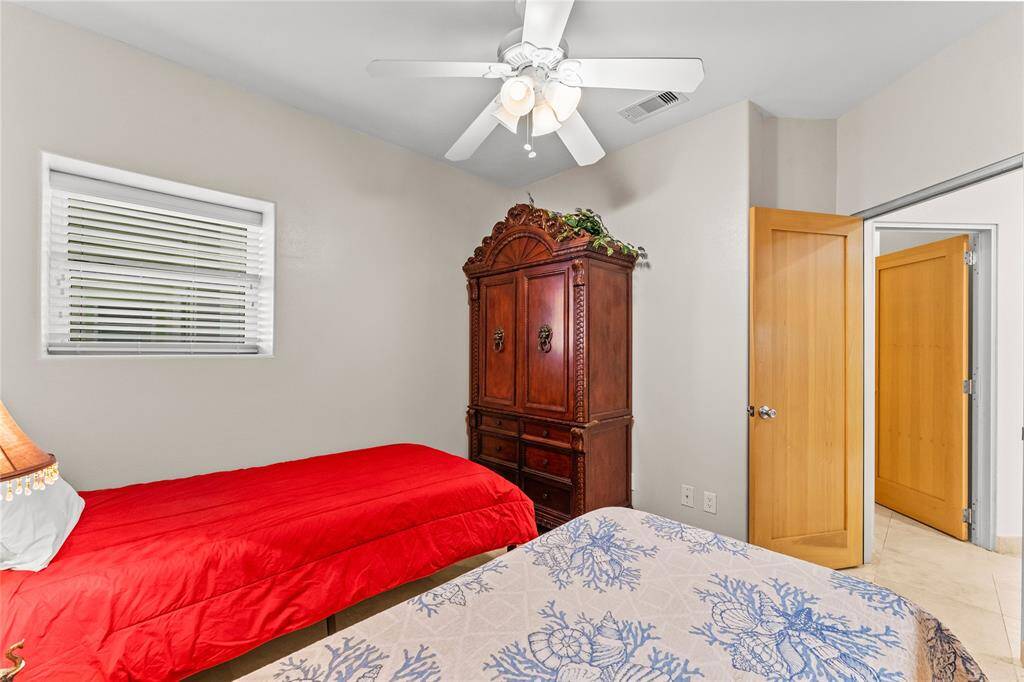 This cozy bedroom features neutral walls, a ceiling fan, and a window with blinds for natural light. It includes a wooden armoire and two beds with vibrant bedding, creating a warm and inviting atmosphere.