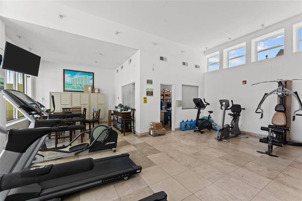 This photo showcases a spacious, well-lit gym with modern exercise equipment including treadmills, stationary bikes, and a multi-gym machine. The room features high ceilings, tiled flooring, and several windows for natural light. Look out over the pool while working out.  Also, mail box area and onsite manager office.