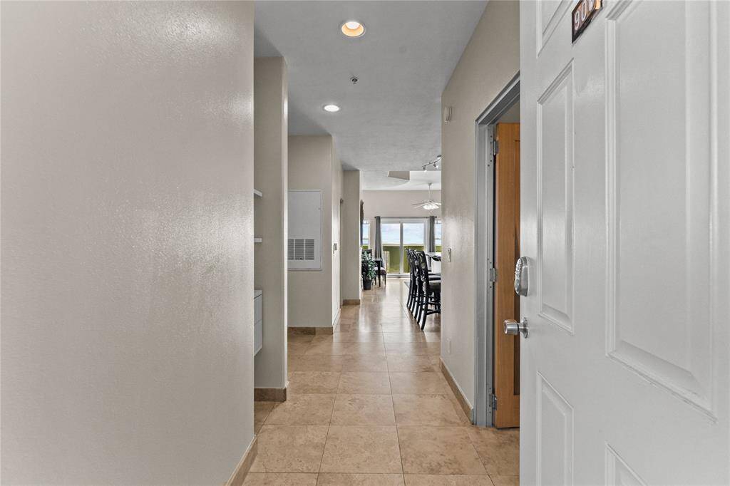 Entering the condo, featuring neutral walls and tiled flooring. The hallway leads to a bright living area with large windows offering a view of the Gulf. Modern lighting fixtures are visible, creating a welcoming and open atmosphere.