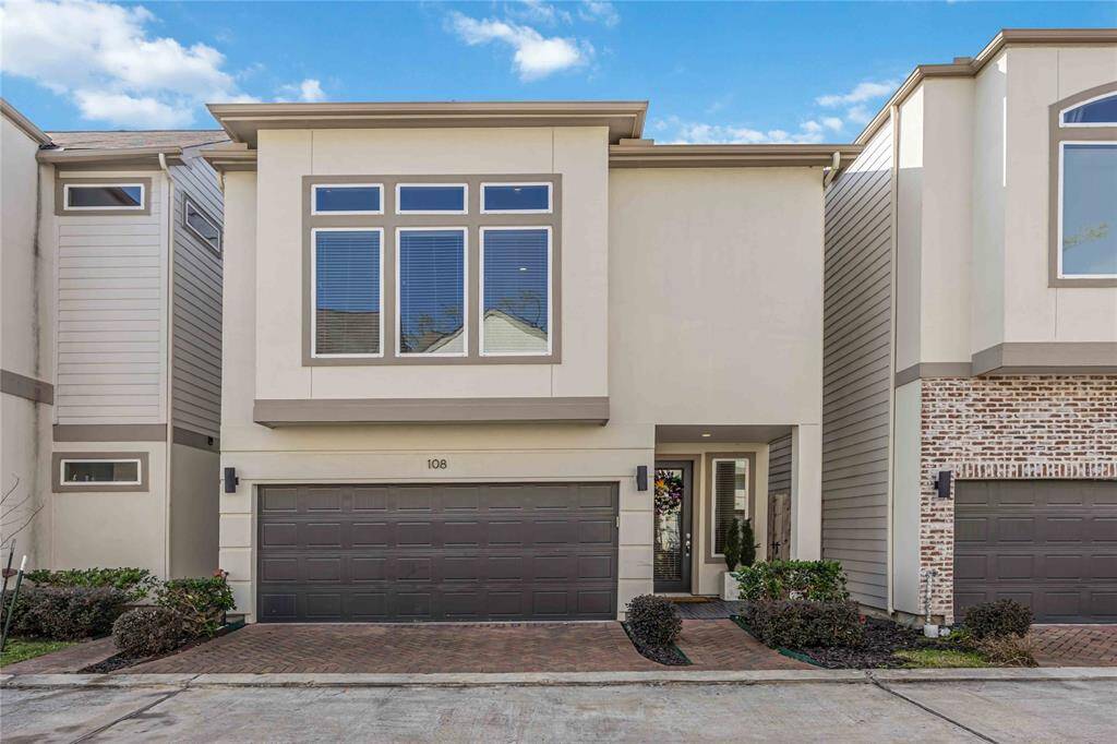 Modern two-story townhouse with a sleek facade, featuring large windows and a two-car garage. The entryway is inviting, with neat landscaping and a clean brick driveway. Ideal for buyers seeking contemporary design.