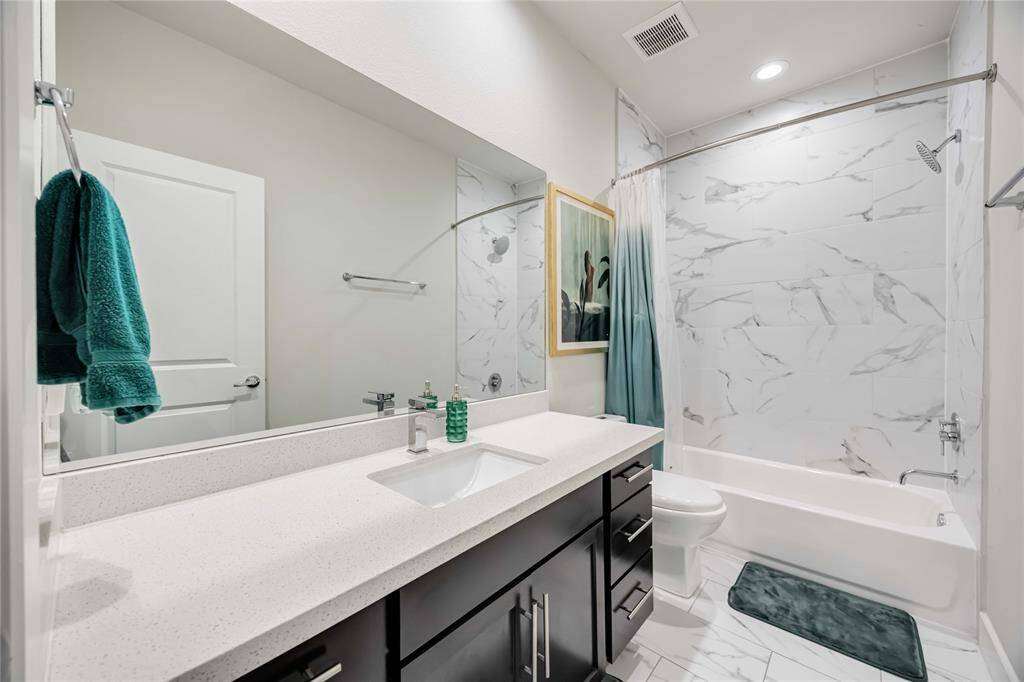 This modern bathroom features a sleek white vanity with ample storage, a large mirror, and a contemporary sink. The shower-tub combo is accented with elegant marble-look tiles, and the space is well-lit with recessed lighting.