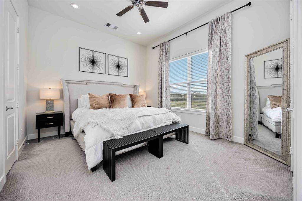 This primary bedroom features a cozy and elegant design with neutral tones, plush bedding, and decorative pillows. It has a large window with a stylish ceiling fan.