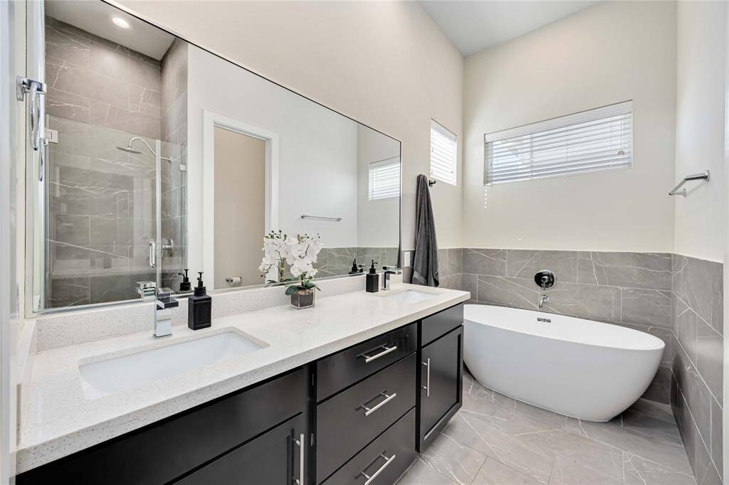 This primary bathroom features a modern design with a sleek double vanity, a large mirror, and a freestanding bathtub. The walk-in shower has a glass door and contemporary tile work. Natural light filters in through high windows, adding to the spacious feel.