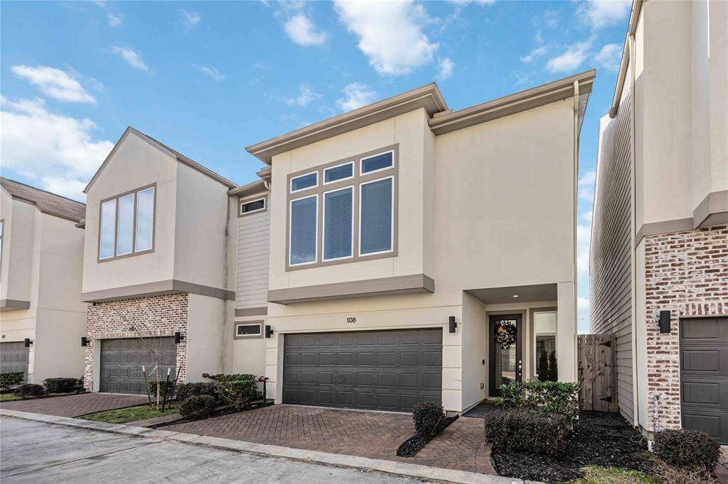 Modern two-story townhouse with a sleek facade, featuring large windows and a two-car garage. The entryway is inviting, with neat landscaping and a clean brick driveway. Ideal for buyers seeking contemporary design.