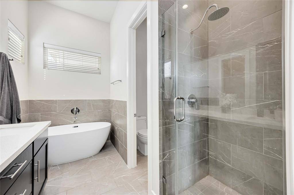 This modern bathroom features a sleek freestanding bathtub, a spacious glass-enclosed shower with elegant tile work, and a contemporary vanity with ample storage. Natural light filters in through a window with blinds, creating a bright and inviting space.
