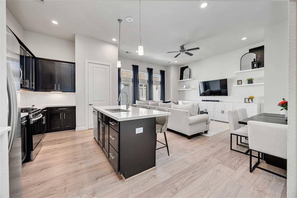 This photo showcases a modern open-concept living space featuring a sleek kitchen with dark cabinets, a central island with seating, and stainless steel appliances. The adjoining living area includes a cozy seating arrangement, built-in shelving, and a wall-mounted TV. Light wood flooring and contemporary lighting create a bright and inviting atmosphere, with ample natural light from large windows.