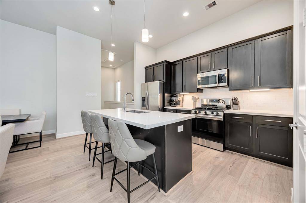 This modern kitchen features sleek dark cabinets, stainless steel appliances, and a spacious island with seating. The open layout and light wood flooring create an inviting atmosphere, perfect for cooking and entertaining.