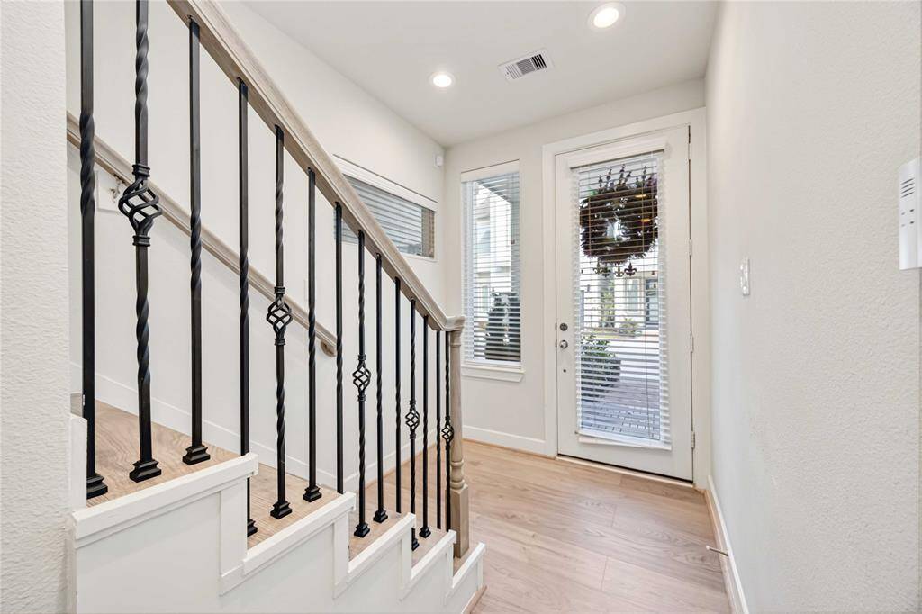 Bright entryway with engineered hardwood floors, featuring a staircase with decorative metal railing. A glass door with blinds leads outside, allowing natural light to fill the space.