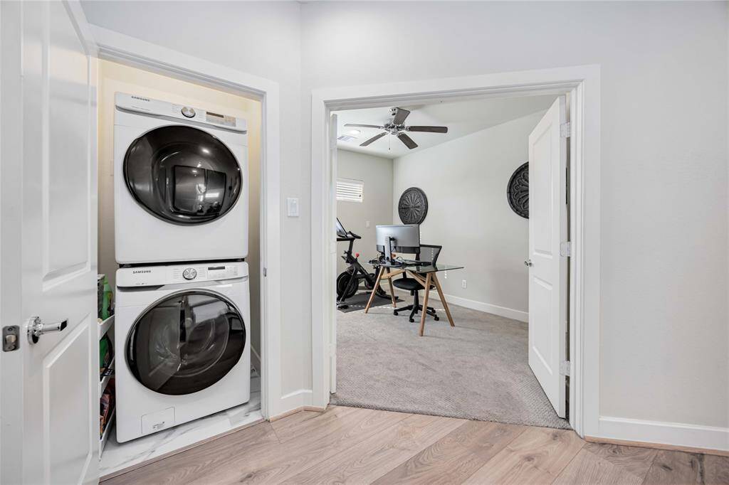 This photo features a compact laundry area with a stacked washer and dryer adjacent to a home office. The office includes a desk, chair, computer setup, exercise bike, and a ceiling fan, offering a functional and versatile space.