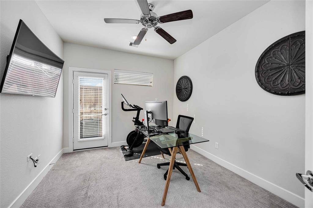 This room features a modern home office setup with a glass desk and computer, complemented by an exercise bike. It has a ceiling fan, natural light from a door with a glass panel, and decorative wall art, creating a versatile space for work or relaxation. This is one of two bedrooms.