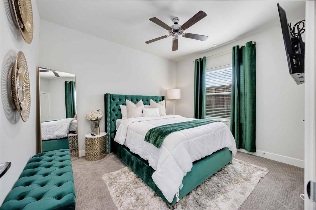 This cozy bedroom features a plush green upholstered bed with matching curtains, a soft white comforter, and a stylish area rug. A ceiling fan and wall-mounted TV add comfort and convenience, while decorative wall accents and a mirror enhance the room's elegance.