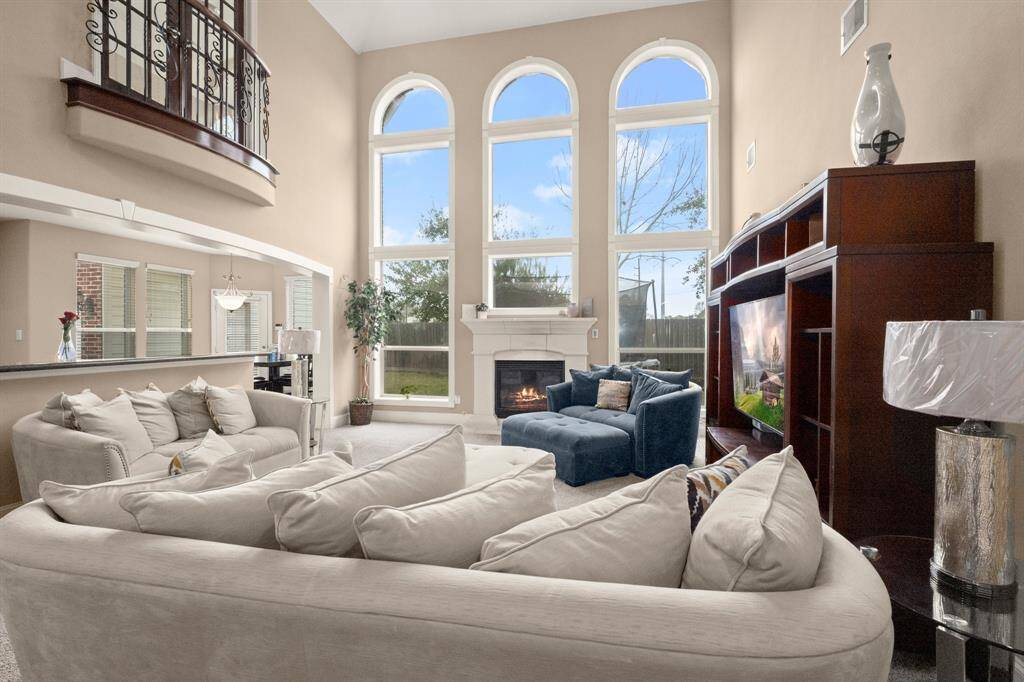 A spacious living room with high ceilings and large arched windows, allowing plenty of natural light. It features a cozy seating area with a plush sectional sofa, a fireplace, and a built-in entertainment center. The open layout connects to the kitchen, creating a welcoming and airy atmosphere.