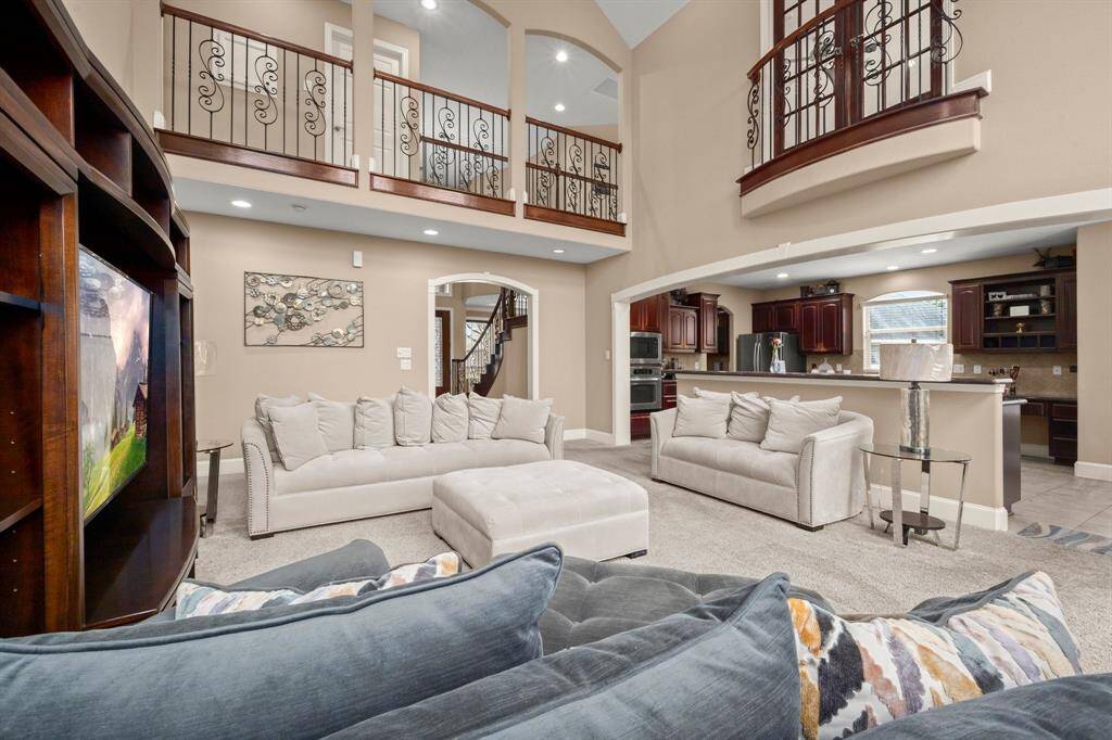 This spacious living area features high ceilings and an open floor plan with elegant wrought-iron railings on the upper level. The room is well-lit and includes comfortable seating, a large entertainment center, and easy access to the modern kitchen with dark wood cabinetry. Perfect for family gatherings and entertaining.
