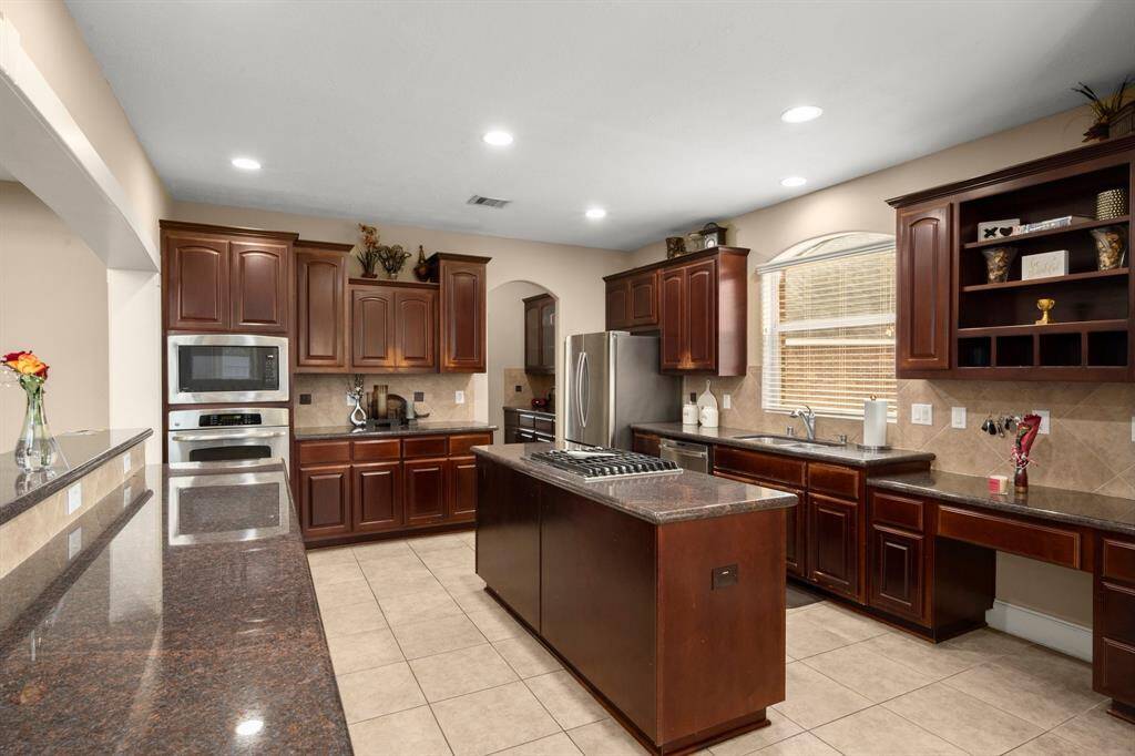 This spacious kitchen features elegant dark wood cabinetry, a central island with a cooktop, stainless steel appliances, and ample counter space. The room is well-lit with recessed lighting and includes a large window for natural light.