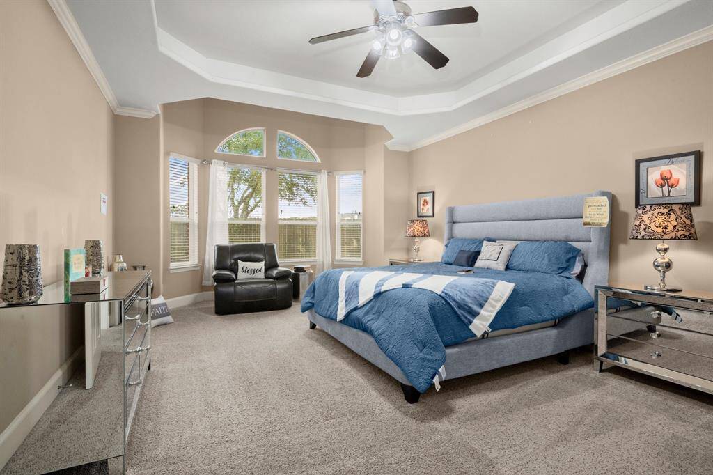 This spacious bedroom features a cozy carpeted floor, a large bed with stylish blue bedding, and a ceiling fan The Bay window offers additional 46 sq ft of sitting area to relax and enjoy mornings or late night talks.  The room is well-lit with natural light from a set of large windows, complemented by elegant decor and mirrored furniture, creating a comfortable and inviting atmosphere.