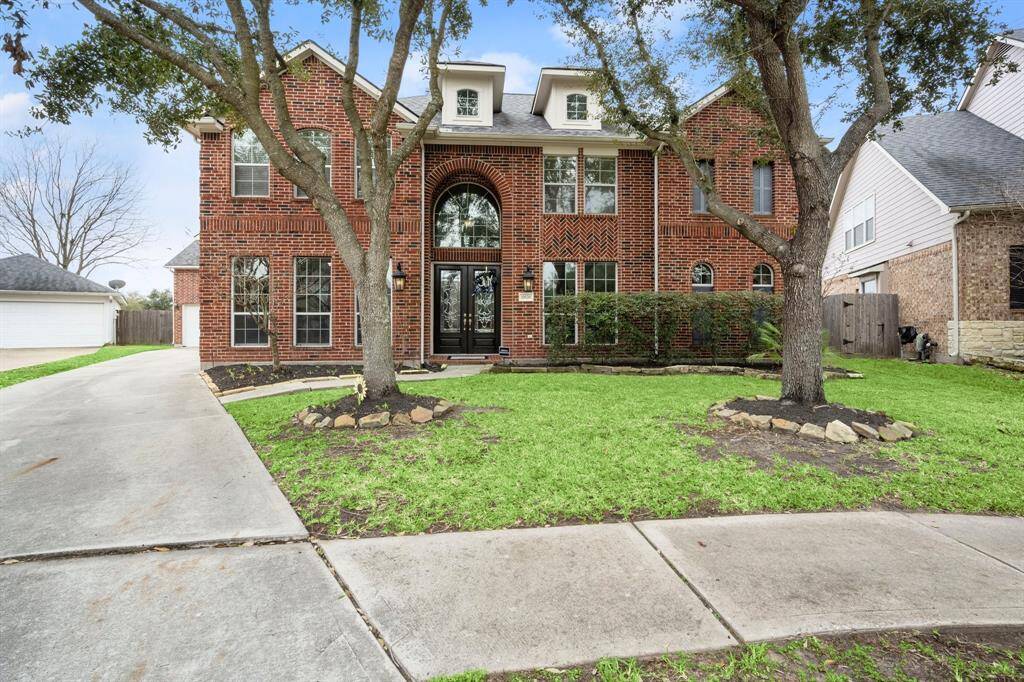 This is a two-story brick home with a classic facade, featuring 8" French double door entry and Large windows Capturing the light throughout the home. It has a well-maintained front lawn with mature trees and a wide driveway leading to a garage. The house is in a suburban neighborhood setting.