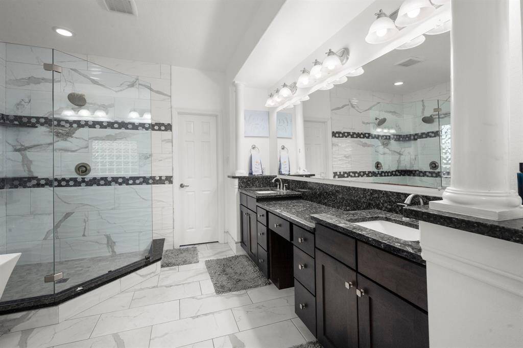 This spacious bathroom features a sleek double vanity with granite countertops, a large mirror, elegant lighting, and a luxurious glass-enclosed shower with stylish tile work.
