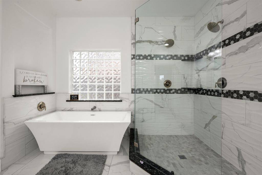 This bathroom features a modern design with a spacious glass shower, elegant marble-patterned tiles, and a sleek freestanding bathtub. Natural light enters through a glass block window, enhancing the bright and airy feel.