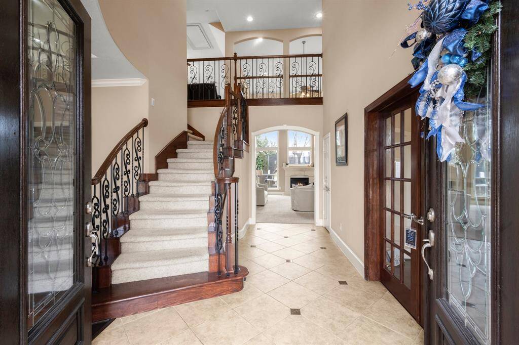 This inviting entryway features elegant tile flooring, a grand staircase with wrought iron railings, and high ceilings. The space opens up to a cozy living area with a fireplace, and there's a stylish glass door adorned with festive decorations. Perfect for welcoming guests and creating a warm home atmosphere.