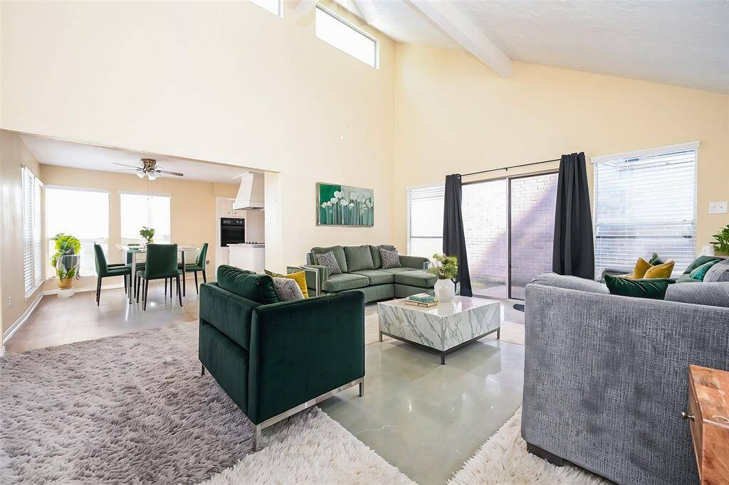 This is a spacious, well-lit living room with high ceilings, large windows, and a sliding glass door leading to an outdoor space. The open layout provides a view into the adjoining kitchen area.