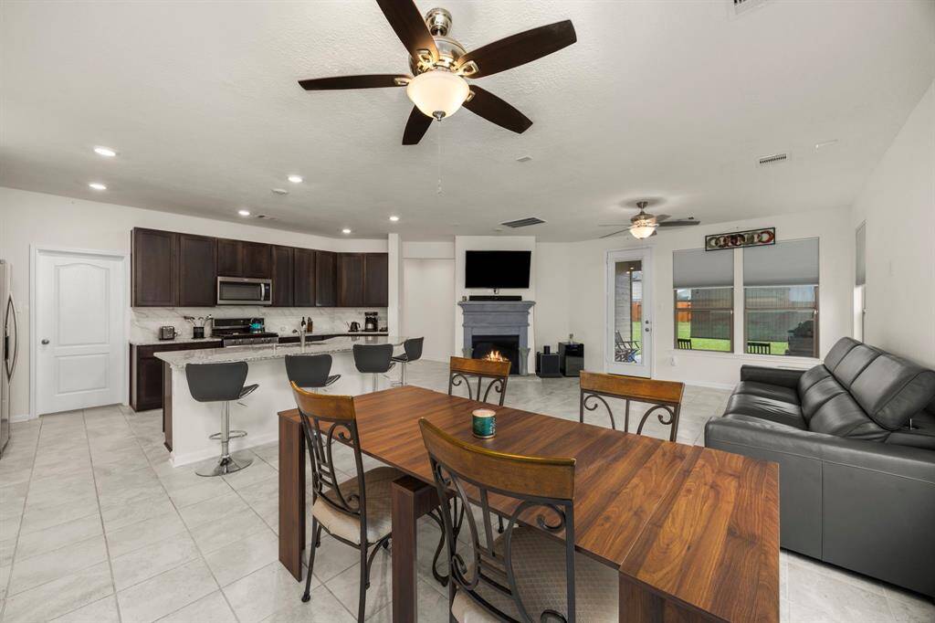 This open-concept living area features a modern kitchen with dark cabinetry, a spacious island with bar seating, and stainless steel appliances. The adjoining living room includes a cozy fireplace, comfortable seating, and large windows offering plenty of natural light. A dining area with a wooden table complements the space, perfect for family gatherings.