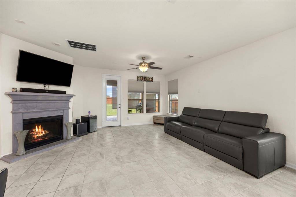 Spacious living room with tile flooring, featuring a cozy fireplace and mounted TV. Large windows and a glass door provide natural light and access to a backyard. Room includes a ceiling fan.