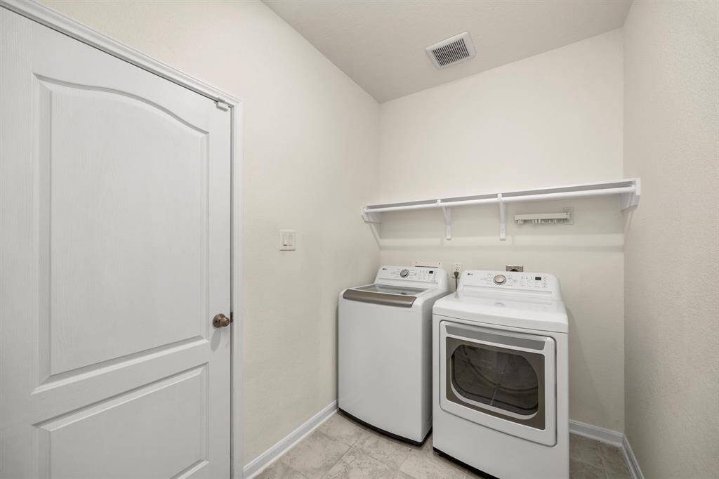 This photo shows a clean, functional laundry room featuring a washer and dryer set, a white door, and a shelf for storage above. The walls and flooring are light, enhancing the room's bright and spacious feel.