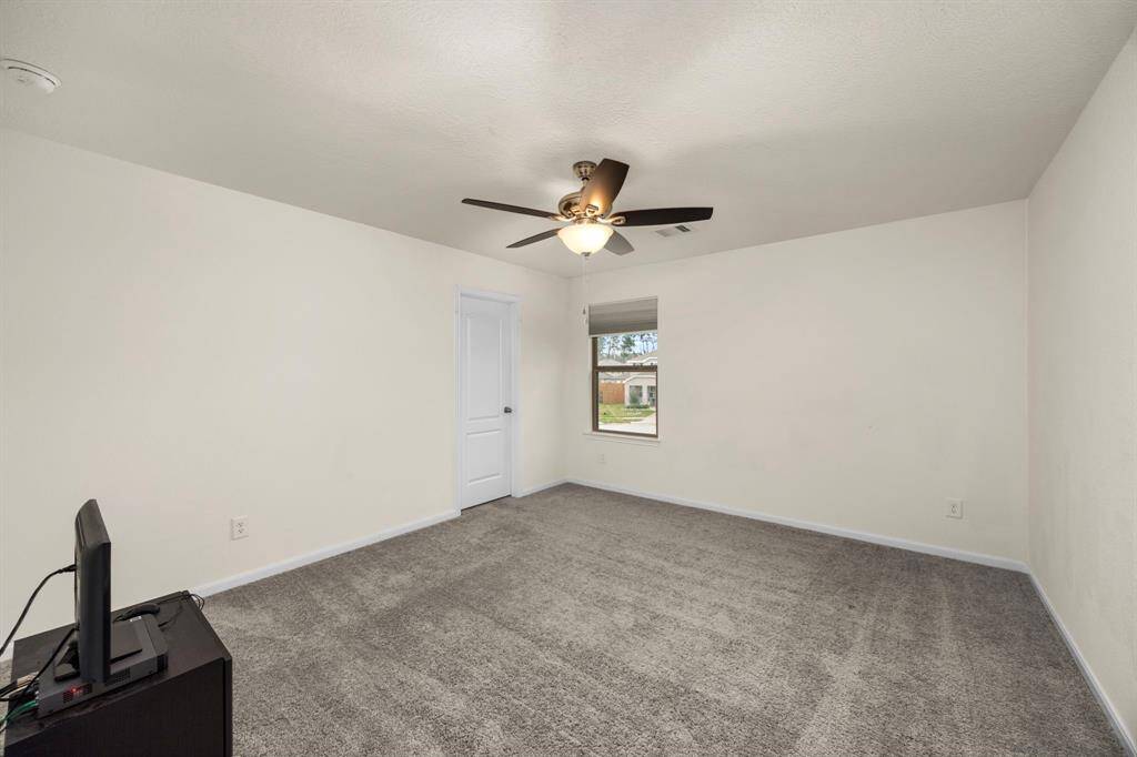 This spacious bedroom features neutral carpeting, a ceiling fan, and a window providing natural light. It includes a door for privacy and ample wall space for customization.