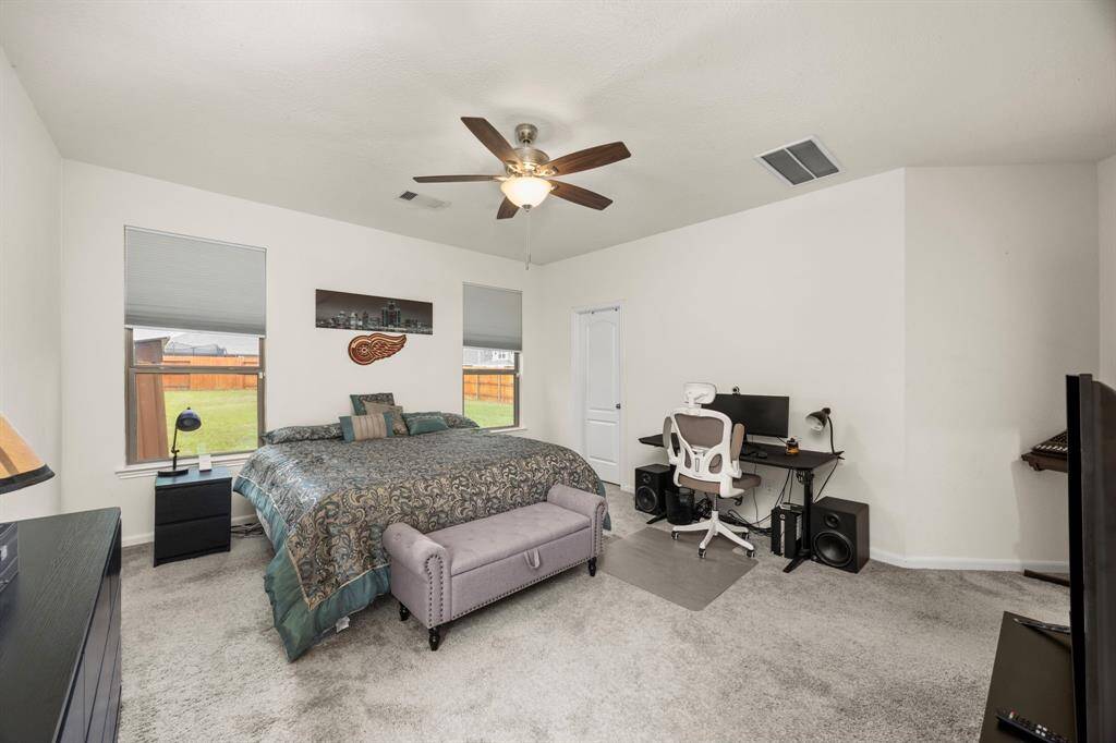 Spacious bedroom with a ceiling fan, featuring a large bed, two windows providing natural light, and a cozy carpeted floor. There's also a functional workspace with a desk and chair, perfect for a home office setup.