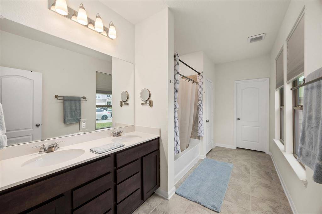 This bathroom features a clean, modern design with dual sinks, dark wood cabinetry, and ample lighting. It includes a shower-tub combo with a curtain, tiled floor, and natural light from multiple windows.