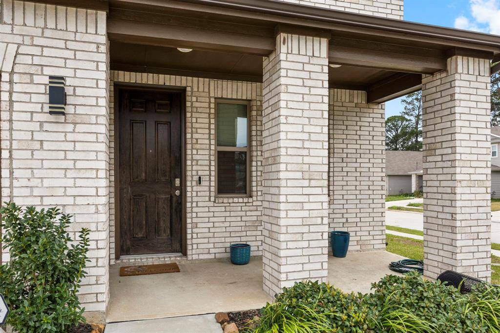 This photo shows the front porch of a home with a covered entryway, featuring light brickwork and a sturdy wooden door. The area is complemented by plants and outdoor lighting, offering a welcoming and well-maintained appearance.