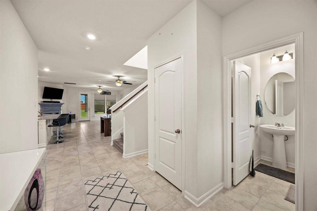 This photo showcases a bright and spacious entryway with tiled floors, leading to an open living area. There's a modern half-bathroom with a pedestal sink and a staircase to the upper level. The space is well-lit with recessed and ceiling lighting, and a door leads to the backyard.