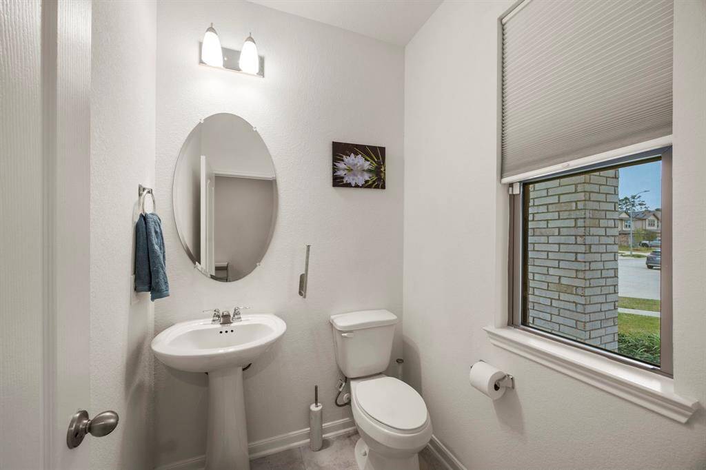 This is a bright, modern half-bathroom featuring a pedestal sink, toilet, oval mirror, and a window with a view. It has neutral tones and simple decor, creating a clean and welcoming space.